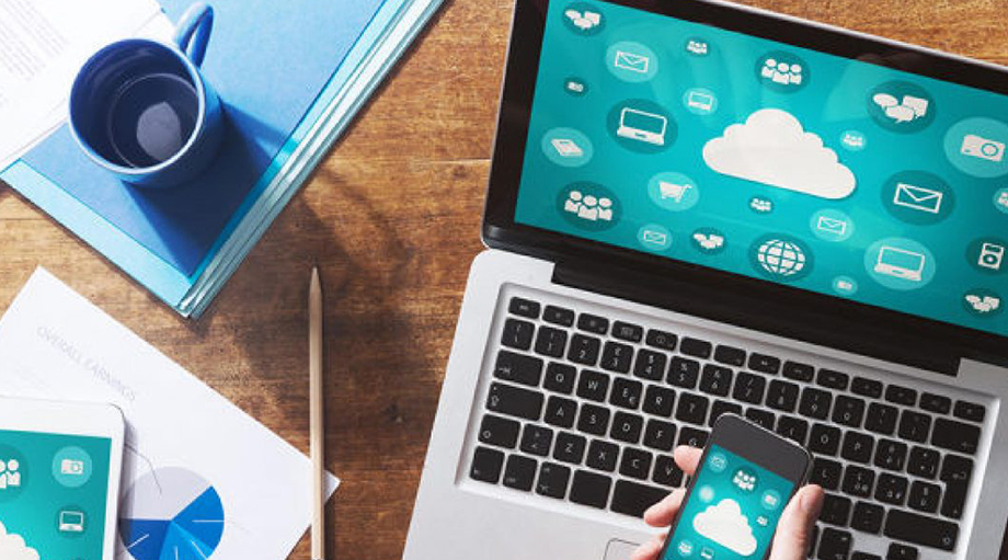 A laptop and smartphone display cloud technology icons on a wooden desk with charts, a pencil, and a blue mug, conveying a digital, tech-focused vibe.
