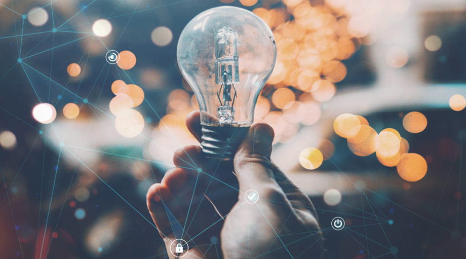 A hand holds a light bulb against a blurred background of warm, glowing bokeh lights.