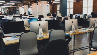 A spacious, modern office with rows of Apple computers on wooden desks. People are working in the background, creating a focused and busy atmosphere.