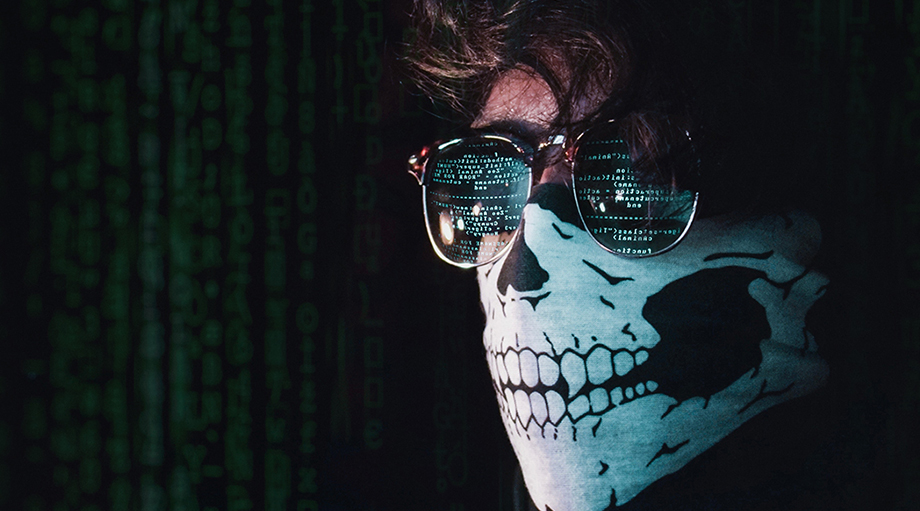 A person wearing sunglasses with digital code reflected in the lenses, their face covered by a bandana featuring a skull design, set against a code-filled background.