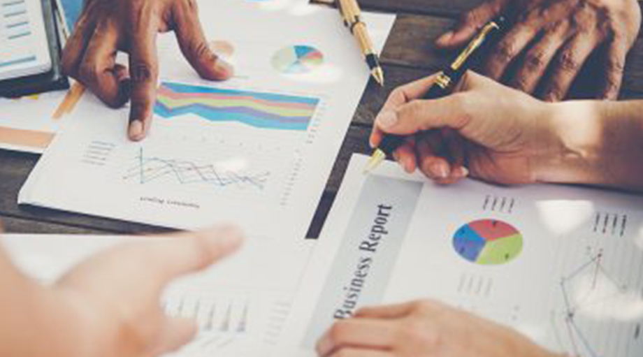 Hands of diverse individuals point at and discuss colorful charts and graphs on business reports spread out on a wooden table.