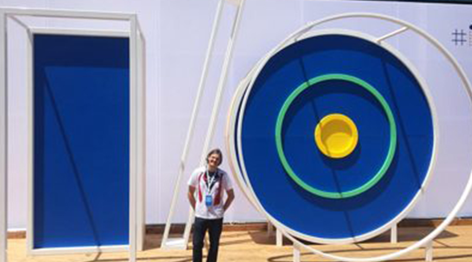 A person smiling stands beside large blue and white structures resembling geometric shapes.