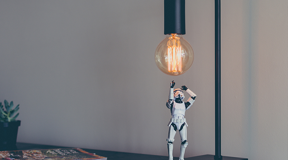 A small stormtrooper figurine reaches up toward a large, glowing vintage bulb on a sleek desk lamp.