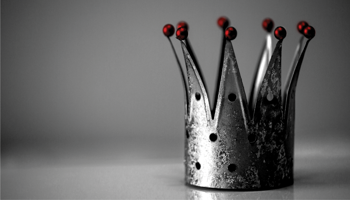 A textured silver crown with five pointed tips, each adorned with a small red sphere, sits on a reflective surface. The background is a smooth gray gradient.