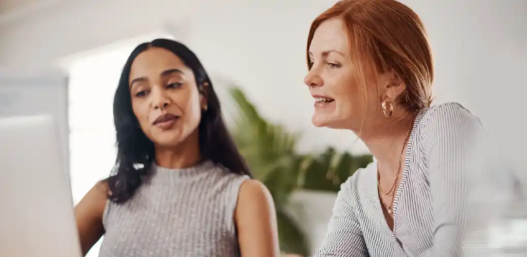 Two women sit together in an office setting, focused on a computer screen. One points, while the other listens intently. The scene feels collaborative.