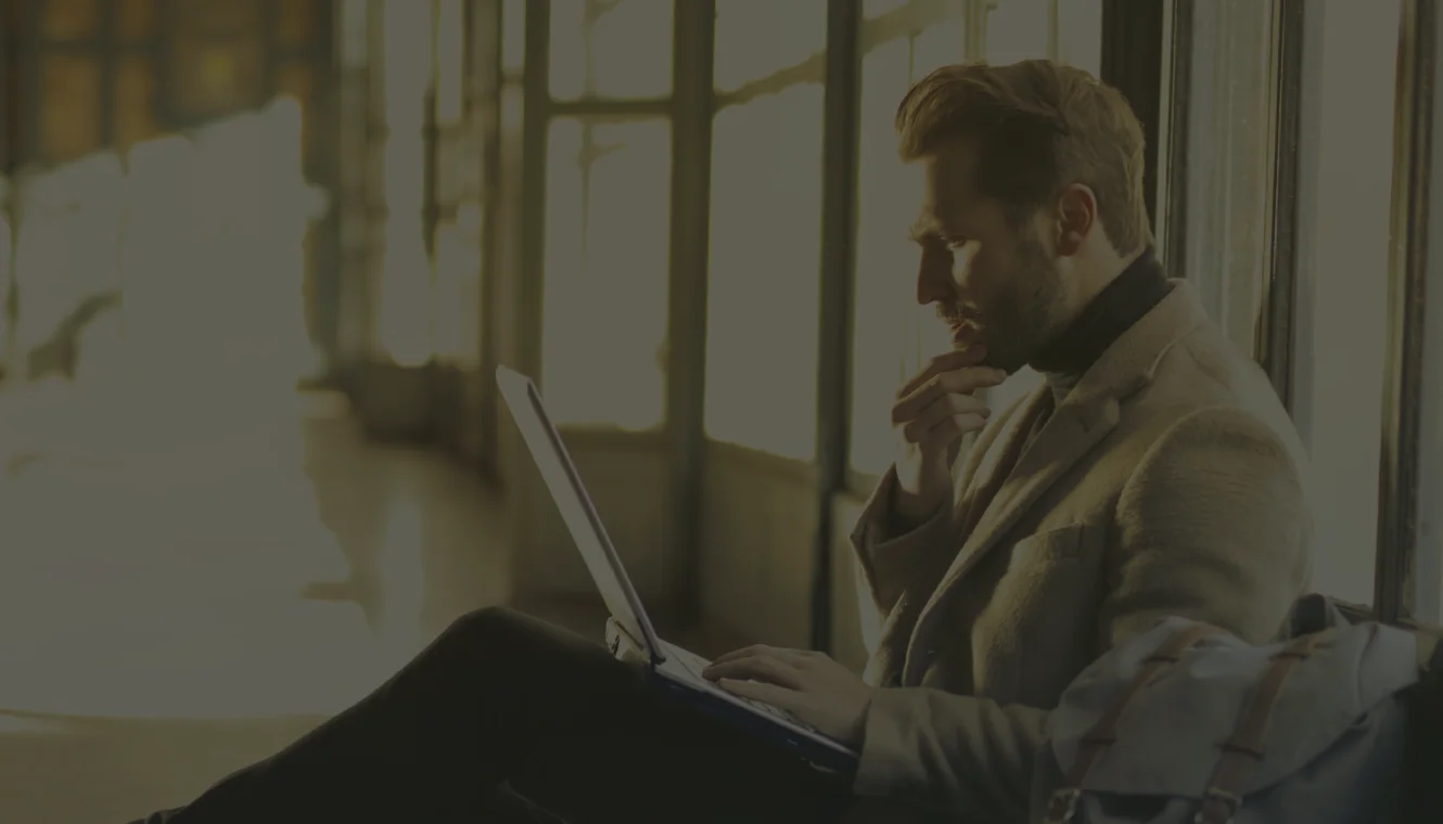A man in a coat sits focused, using a laptop on his lap in a sunlit room. The soft lighting and thoughtful pose create a contemplative atmosphere.
