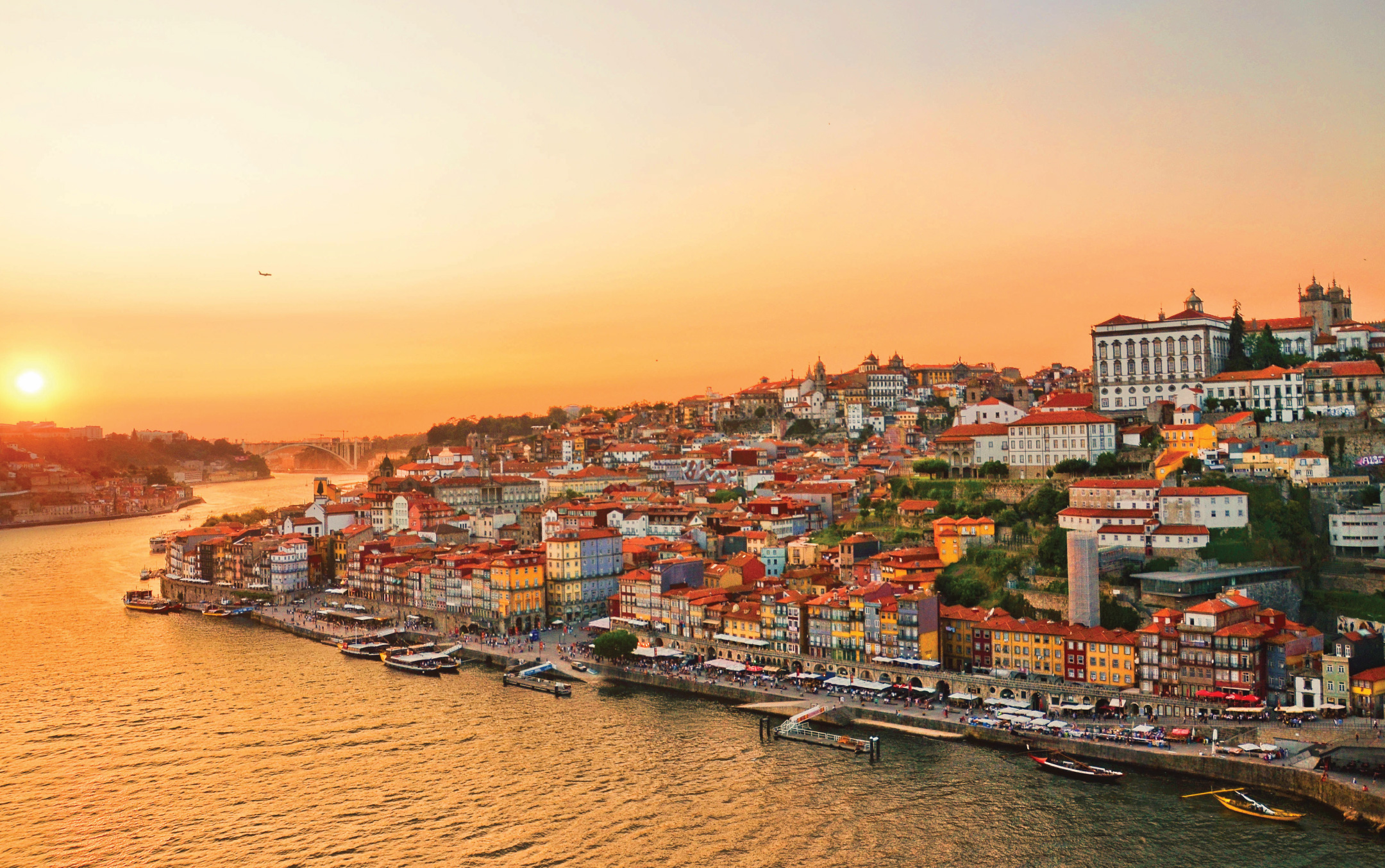 Sunset view of Porto, Portugal, with colorful buildings lining the Douro River. The vibrant sky casts a warm glow, creating a serene, picturesque scene.