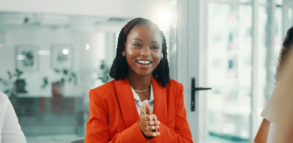 Smiling woman in a bright orange blazer sits in an office environment, clapping hands. Soft lighting creates a warm, positive atmosphere.