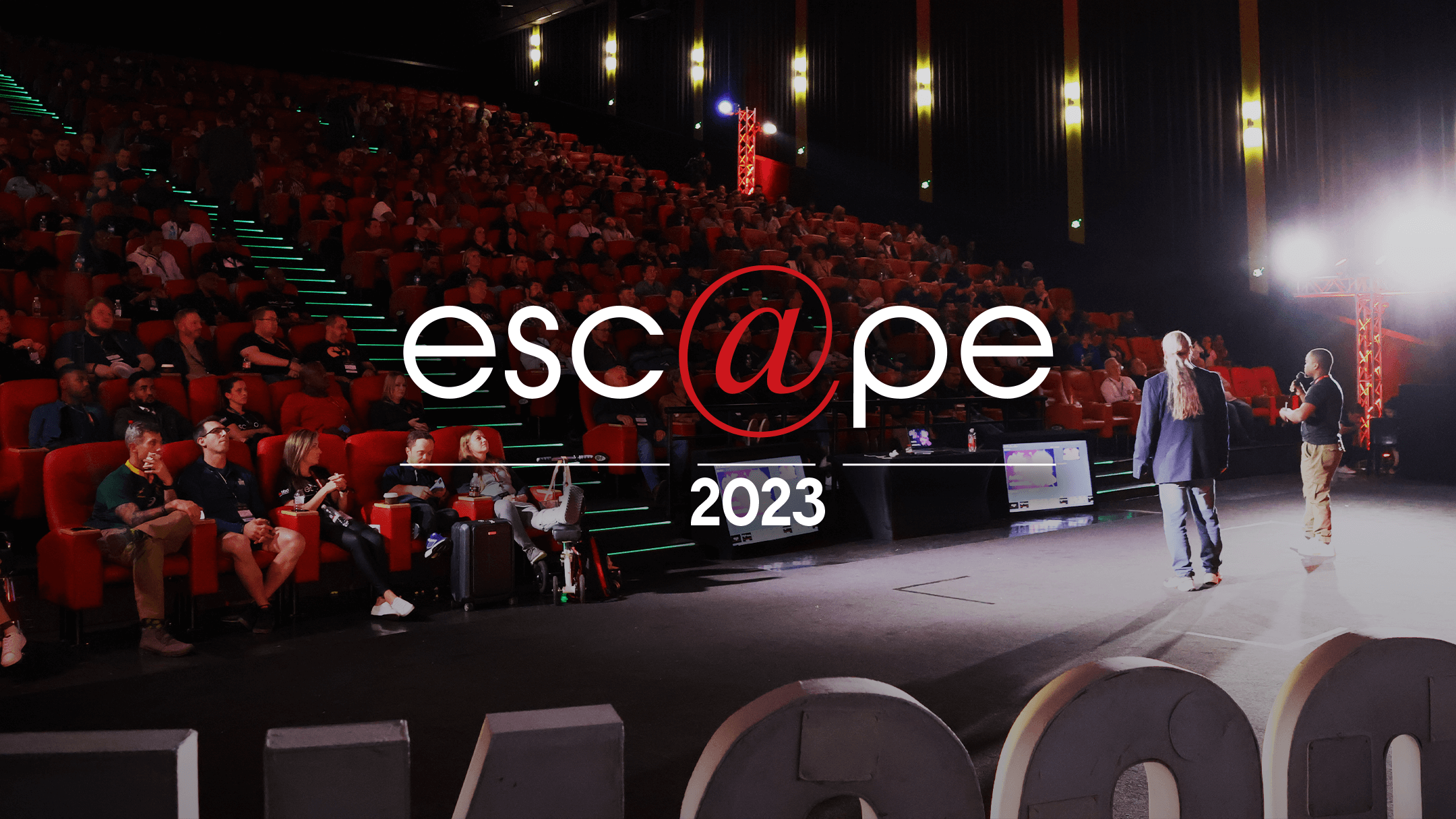 Audience seated in a large theater, dimly lit with red chairs. On stage, two presenters engage the crowd. Text overlay reads "esc@pe 2023."