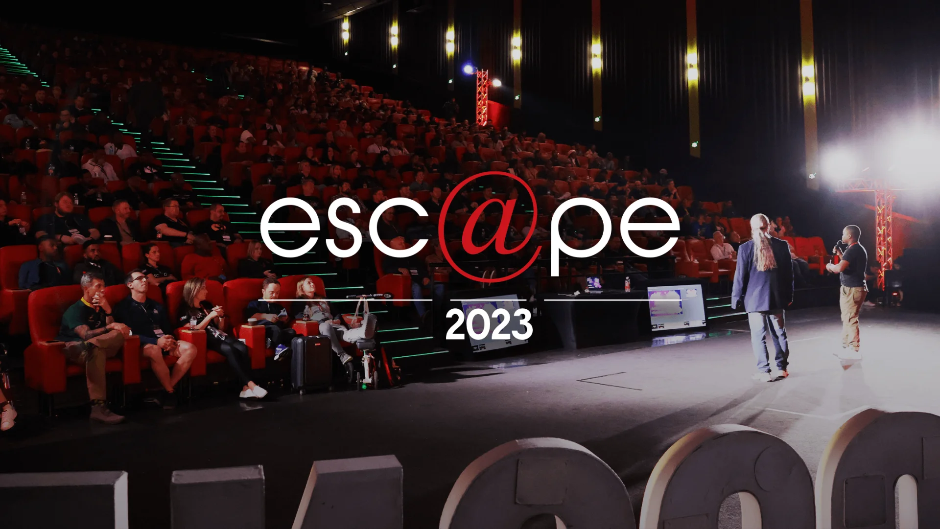 Audience seated in a large theater, dimly lit with red chairs. On stage, two presenters engage the crowd. Text overlay reads "esc@pe 2023."