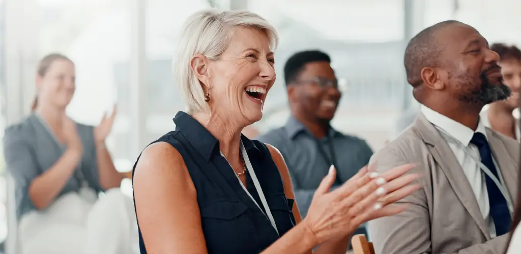 A group of people sit in a bright room, smiling and clapping. A woman in the foreground laughs, creating a joyful and engaging atmosphere.