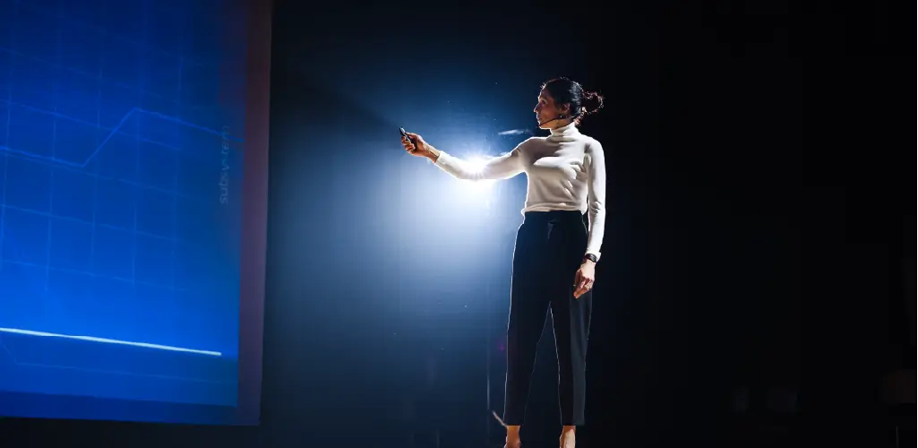 A woman stands on stage pointing at a large blue screen displaying a graph. She wears a headset, projecting a professional and focused demeanor.