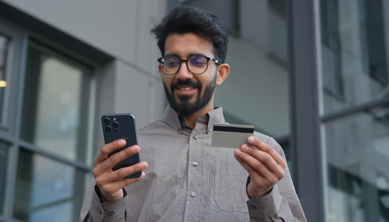 A man with glasses smiles while holding a smartphone and a credit card, standing outside a modern building, conveying a sense of satisfaction and connectivity.