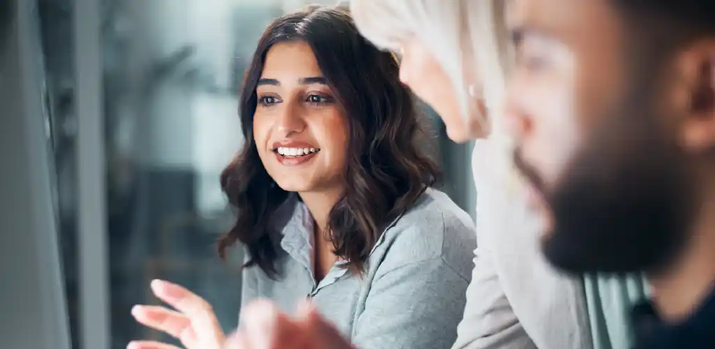 Three people in an office setting engage in a meeting. A woman with brown hair smiles, conveying collaboration and positivity. Blurred background.