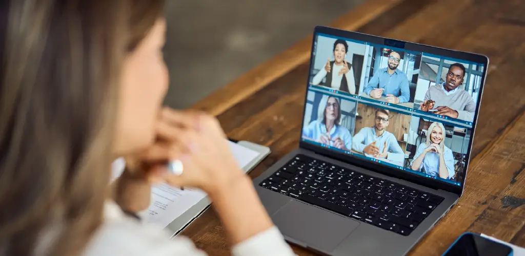A woman participates in a video conference on a laptop, showing six diverse people engaged in discussion. The setting is a wooden table, conveying a professional tone.