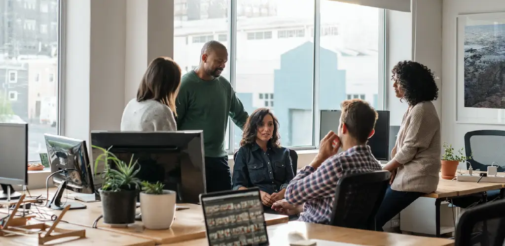 A diverse group of five colleagues is engaged in a discussion in a bright, modern office. Plants and computers are visible, creating a collaborative atmosphere.