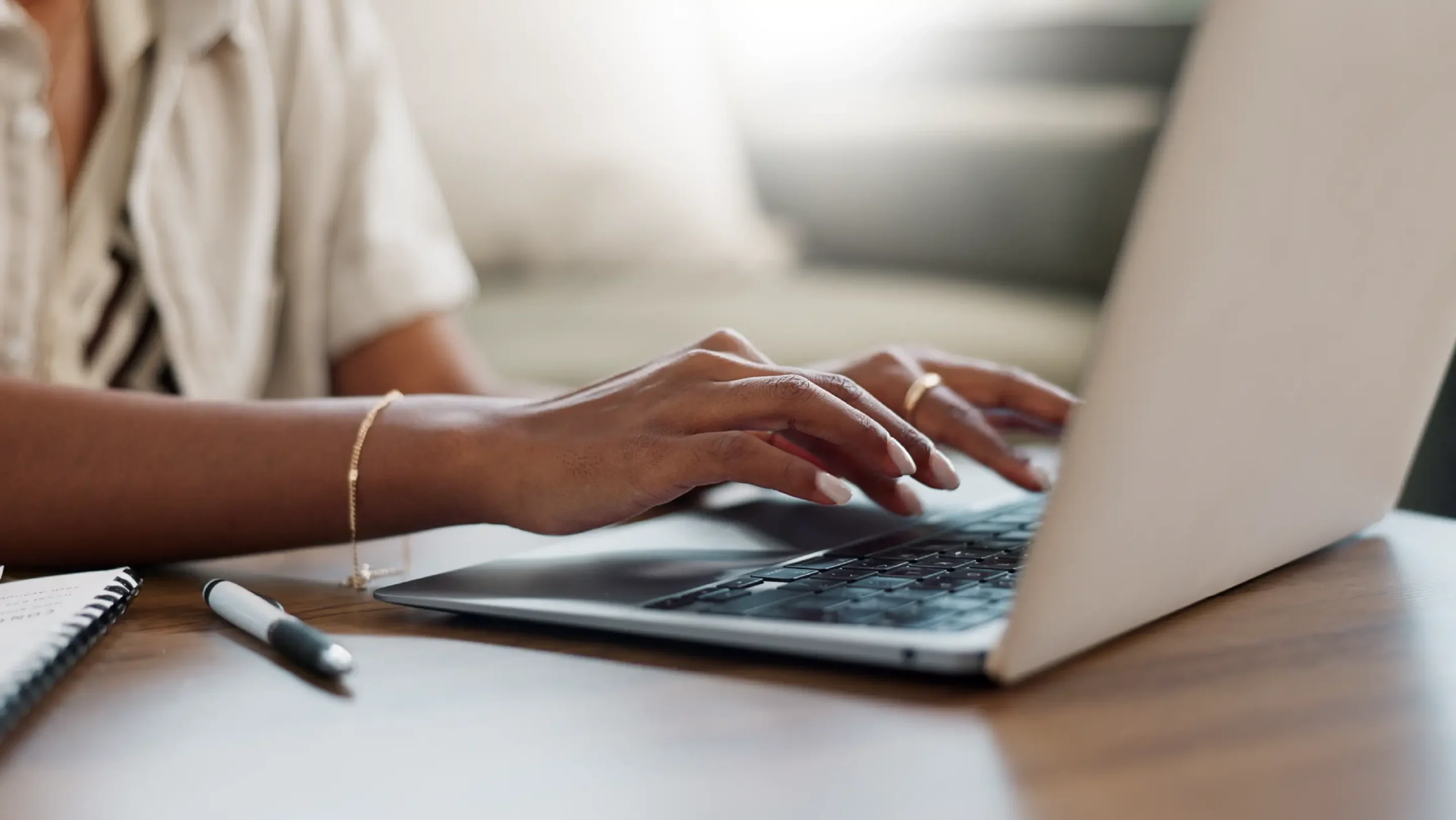Person typing on a laptop at a wooden desk, with a notepad and pen nearby. The scene conveys focus and productivity in a calm setting.