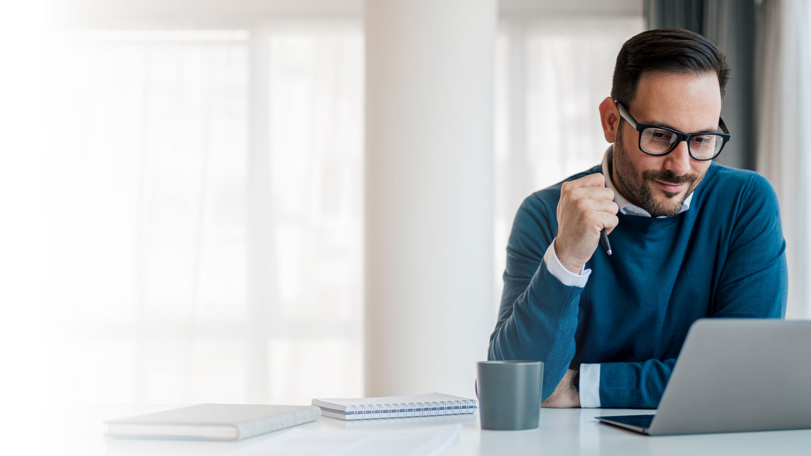 Man in glasses and blue sweater sits at a table, smiling at a laptop. A notebook and coffee cup are nearby. The scene conveys focus and contentment.