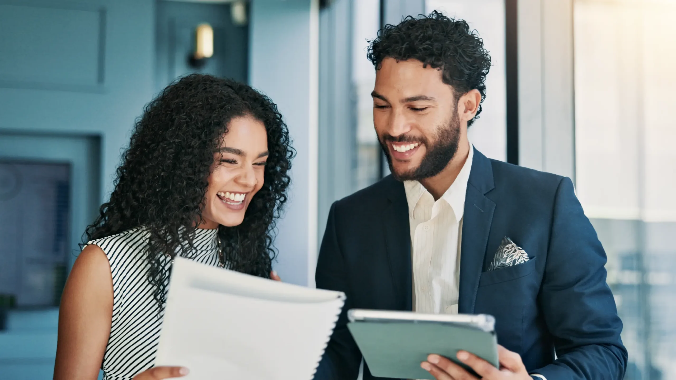 Two business professionals reviewing documents and smiling in a modern office environment.