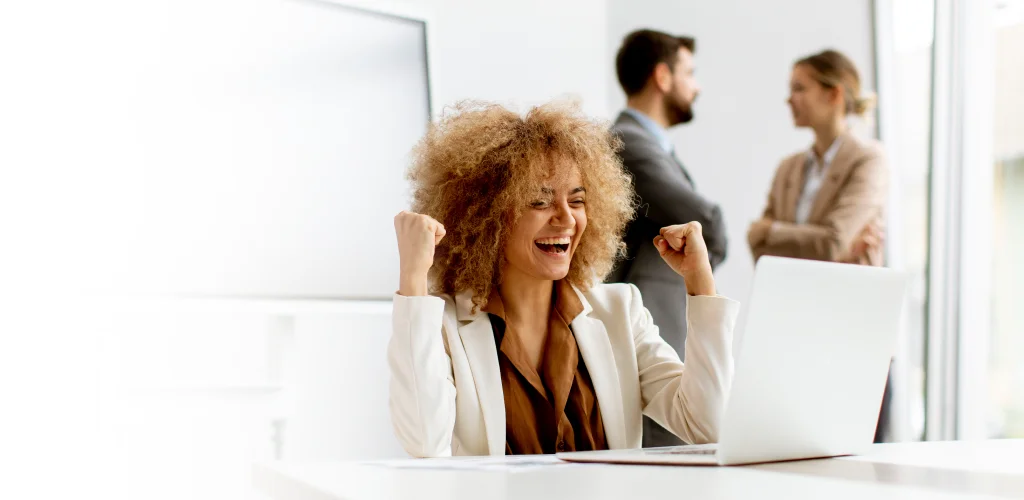 A woman with curly hair celebrates at a desk with a laptop, smiling and raising her fists in joy. In the background, two people converse, slightly out of focus.