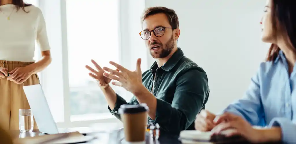 A man in glasses, seated at a desk, gestures expressively during a discussion with two colleagues. A laptop and coffee cup are on the table, conveying a collaborative work environment.