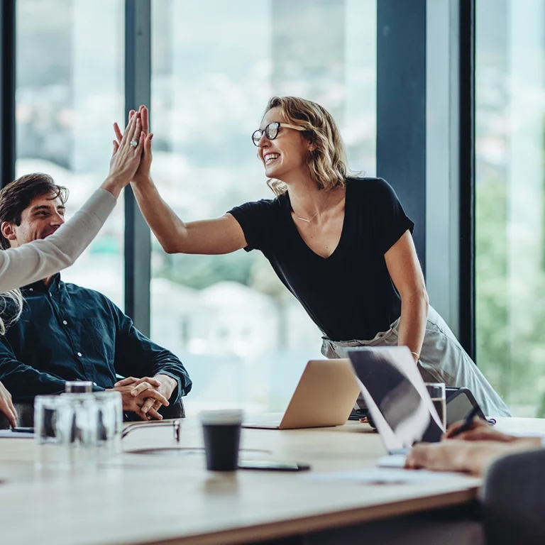Two colleagues giving a high five in a meeting room, symbolising successful teamwork and collaboration.