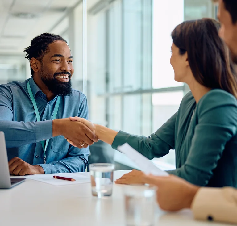 A smiling man in an office shaking hands with a colleague across a meeting table, symbolising partnership and trust.