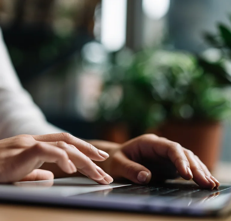 Close-up of hands typing on a laptop with plants in the background, illustrating focus and digital work.