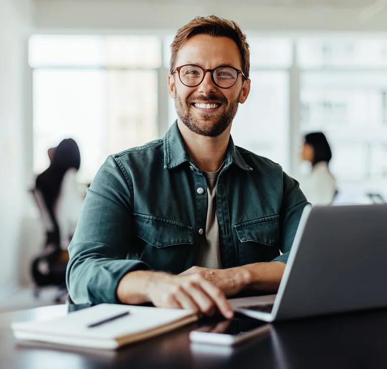 A smiling man sitting at a desk with a laptop and notebook, conveying confidence and a positive work environment.