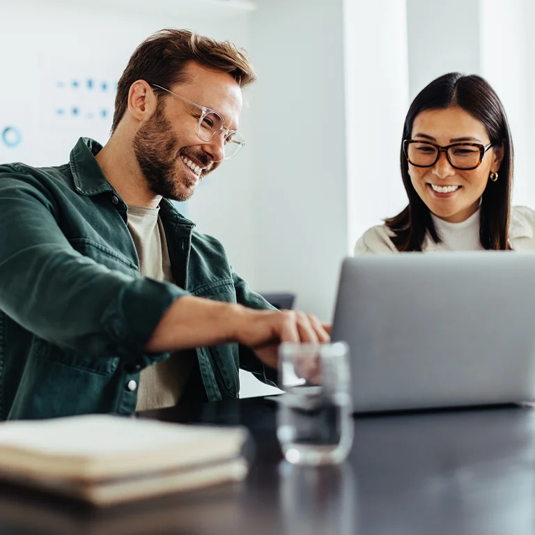 Two smiling colleagues reviewing information on a laptop, representing collaboration in banking technology.