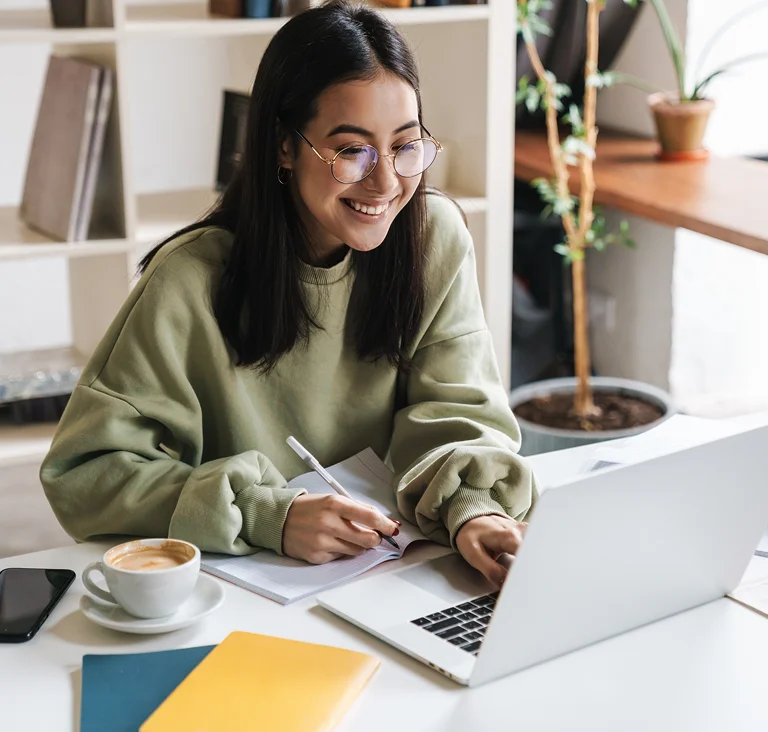 A young woman studying on her laptop with notebooks and coffee on the table, representing online learning.