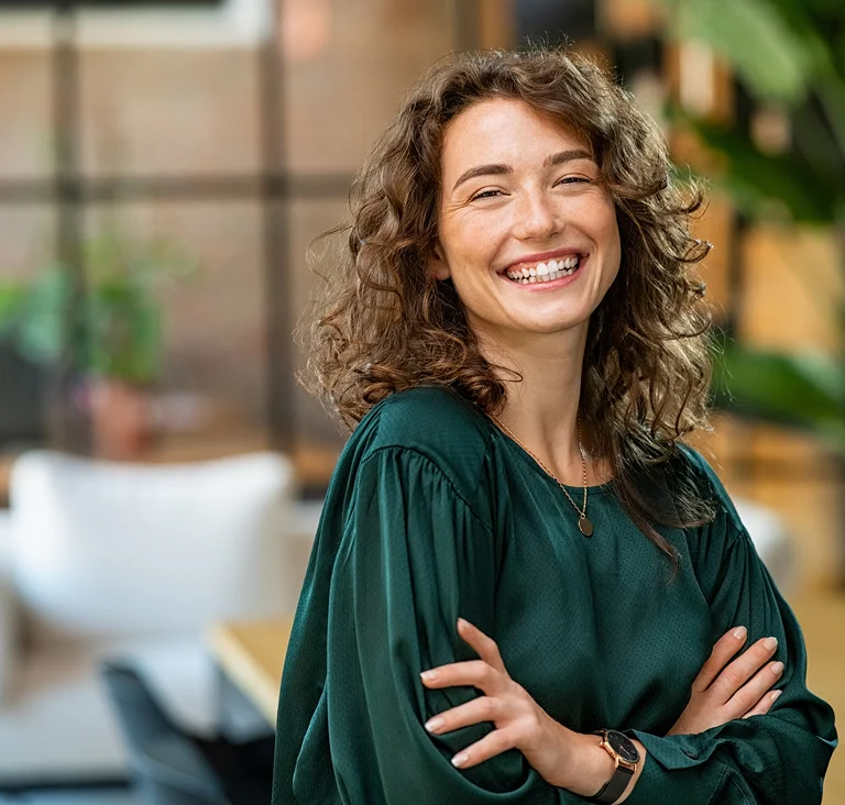 A confident young woman standing in an office with folded arms, smiling warmly, representing leadership and positivity.