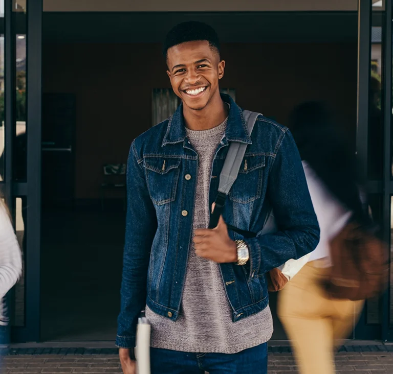 A young man standing outside a building with a backpack, smiling confidently, symbolising student success and opportunity.