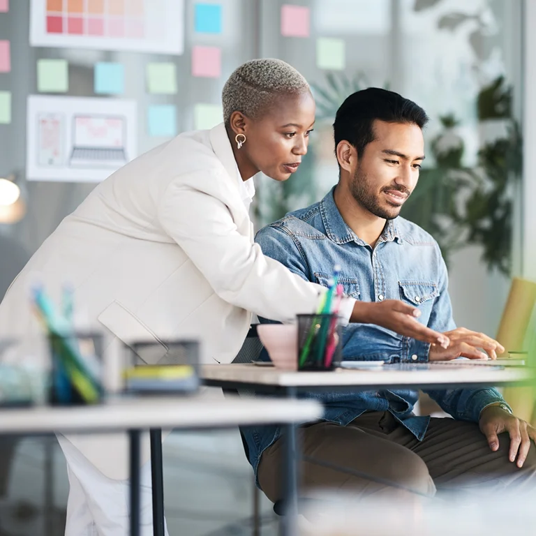 A woman in a white suit guiding a man using a laptop in an office, symbolising cloud migration and project leadership.
