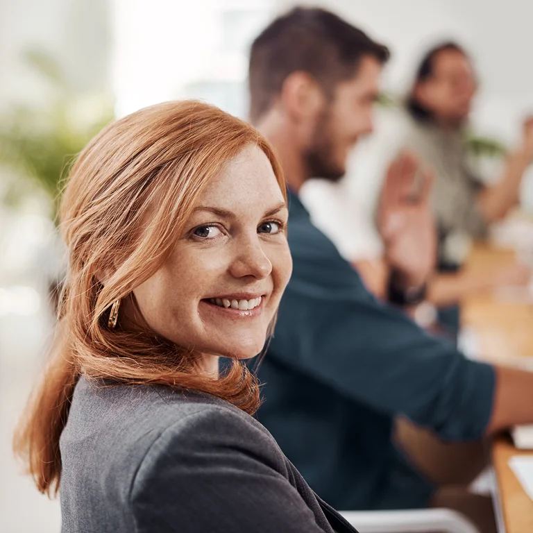 A smiling woman sitting in a meeting, looking back at the camera, representing professionalism and confidence.