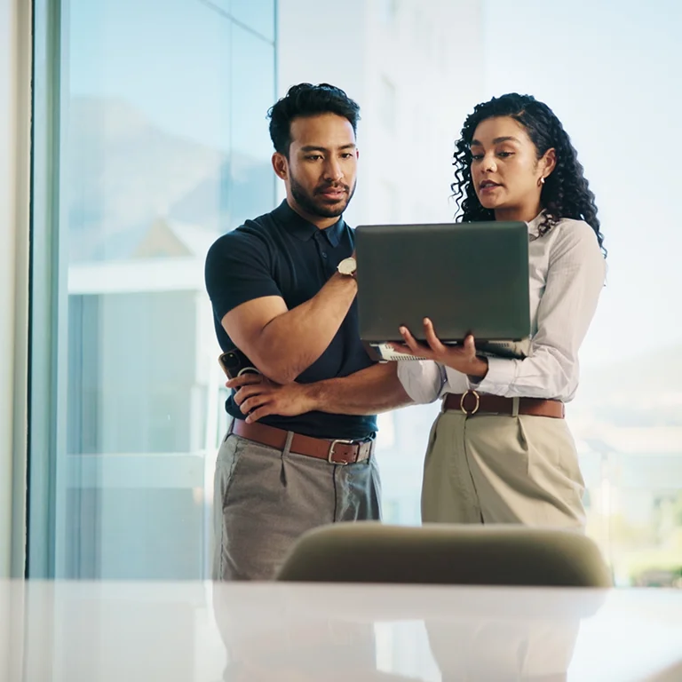 Two professionals standing and reviewing work on a laptop near a window, representing digital transformation in the public sector.