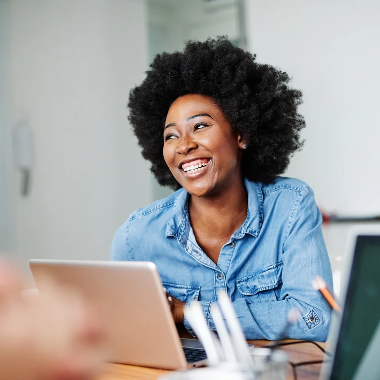A smiling woman working on a laptop in a bright office, symbolising innovation and connectivity in telecommunications.
