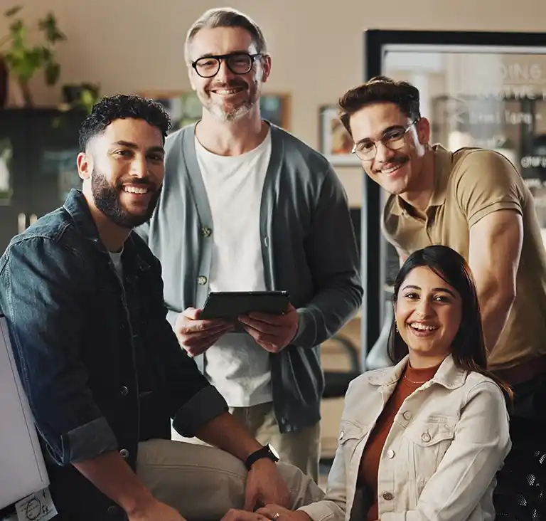 Four people are gathered in an office setting, smiling warmly. A gray-haired man holds a tablet, while the others sit and stand around him, conveying teamwork.