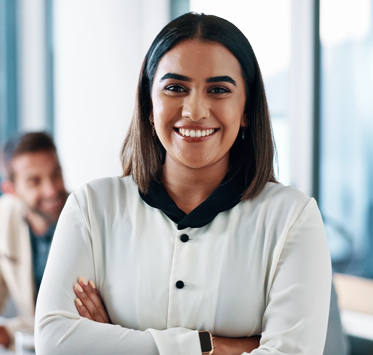 A woman in a white blouse with black buttons stands confidently with arms crossed, smiling warmly. Blurred office background. Positive and professional tone.