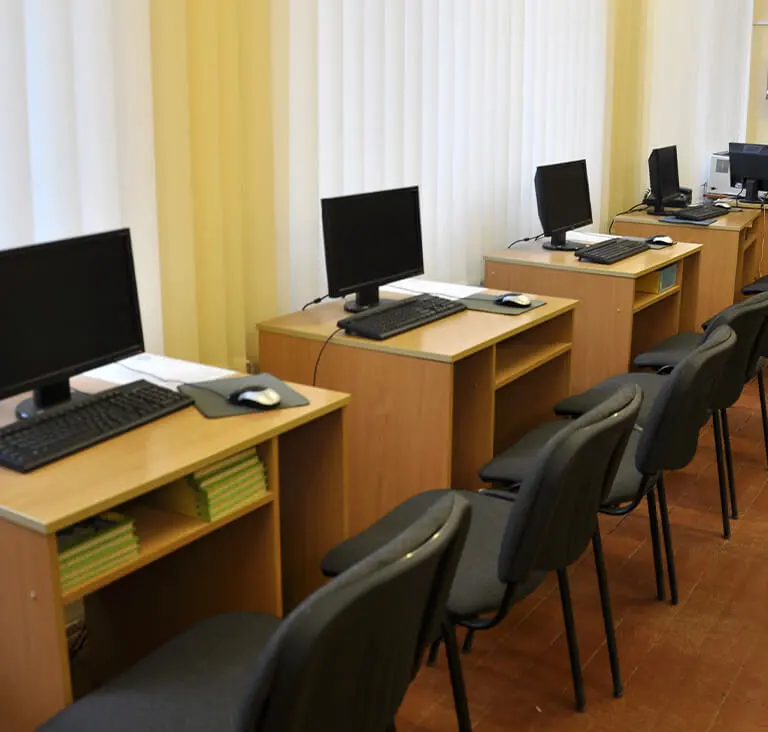 A row of empty computer desks with black monitors and keyboards on light brown wooden tables. Office chairs are neatly aligned, creating a tidy, orderly atmosphere.
