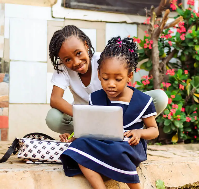 Two children are seated outdoors, focused on a laptop. One smiles warmly while the other is engaged with the screen. Colorful flowers bloom nearby.