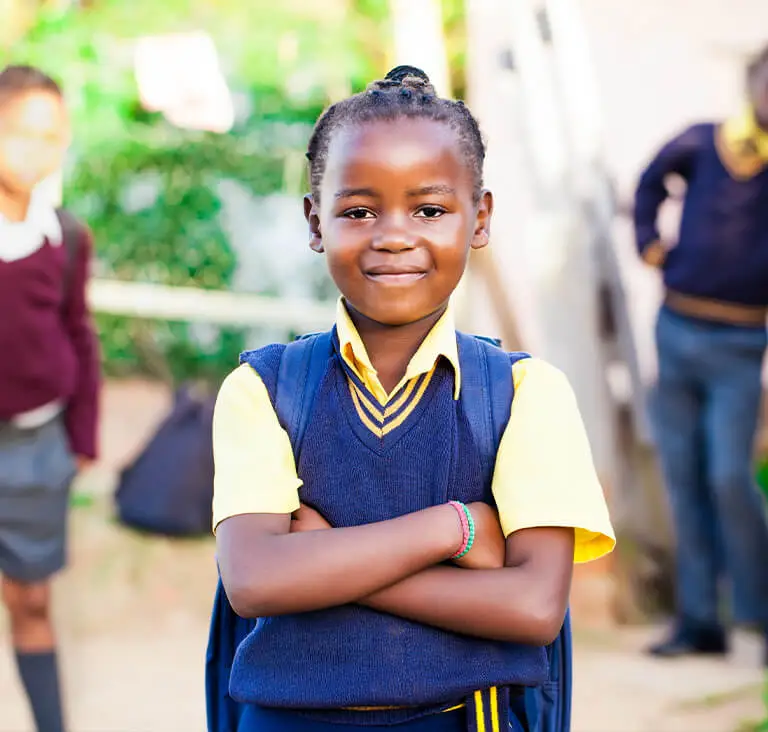 A confident young girl in a yellow shirt and navy vest stands with folded arms, smiling outside a school. Her backpack is visible, and other students can be seen in the background.