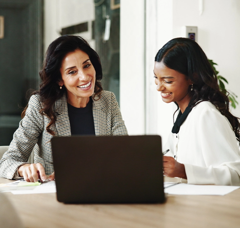 Two women in professional attire smile while discussing a project at a desk with a laptop. The atmosphere is friendly and collaborative.