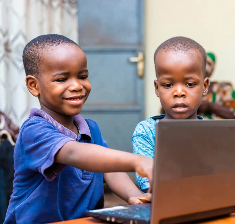 Two young boys are sitting together at a table, engaged with a laptop. The boy on the left smiles while pointing at the screen, suggesting learning or play.
