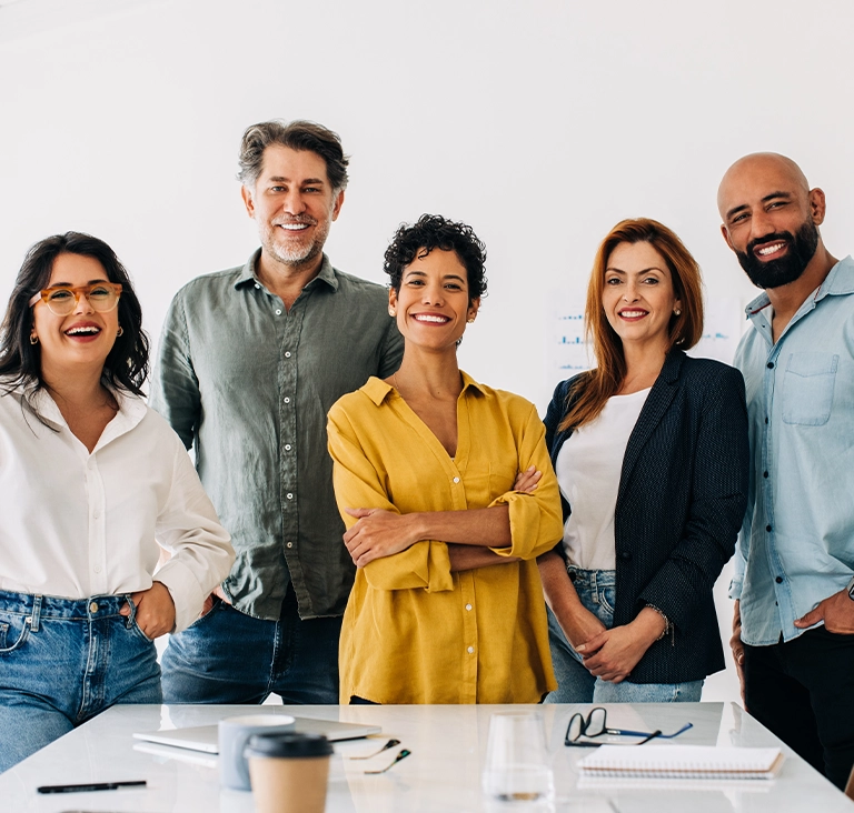 A diverse group of five people stands smiling in an office setting. They exude confidence and camaraderie, surrounded by desks with notebooks and coffee.