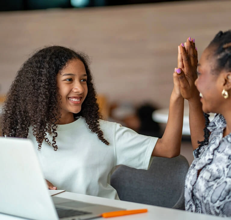 A young girl and a woman share a joyful high-five at a desk with a laptop, conveying success and teamwork. The setting is bright and modern.