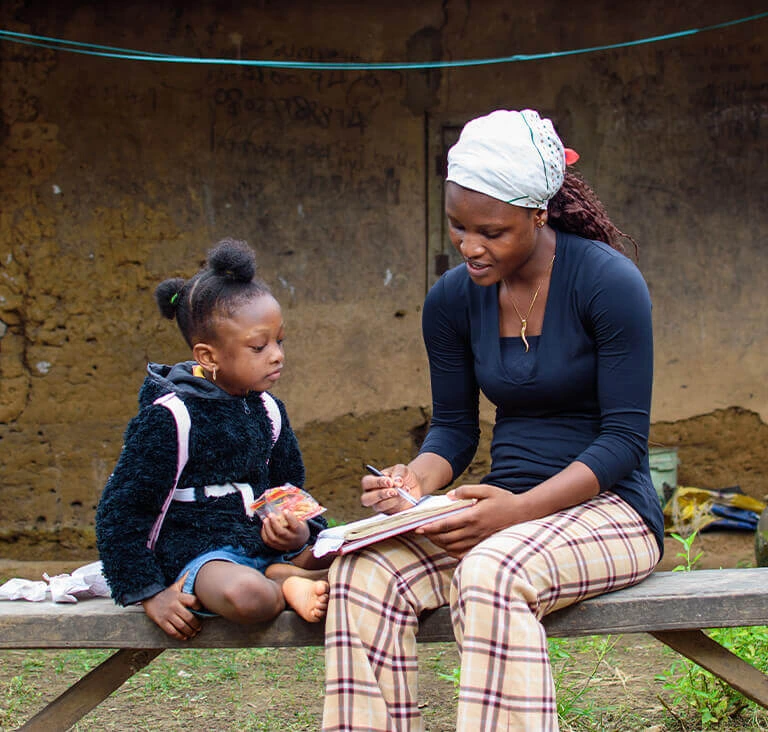 A young woman wearing a headscarf and plaid pants sits on a bench, writing in a notebook. A little girl in a fluffy coat sits beside her, holding a snack. The scene conveys a sense of learning and mentorship outdoors with a rustic background.