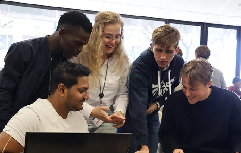 Group of five young adults collaborating around a laptop in a bright room, smiling and engaged, conveying teamwork and a positive atmosphere.