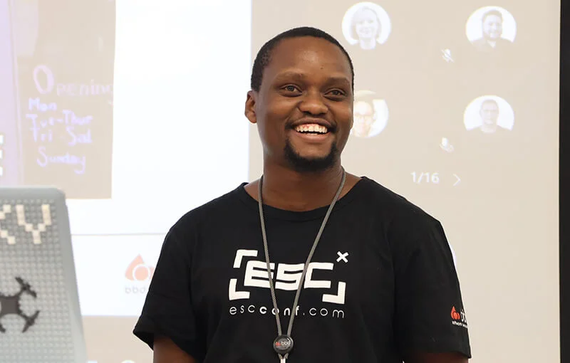 Smiling man wearing a black T-shirt, standing in front of a presentation screen with icons. The atmosphere is upbeat and engaging.