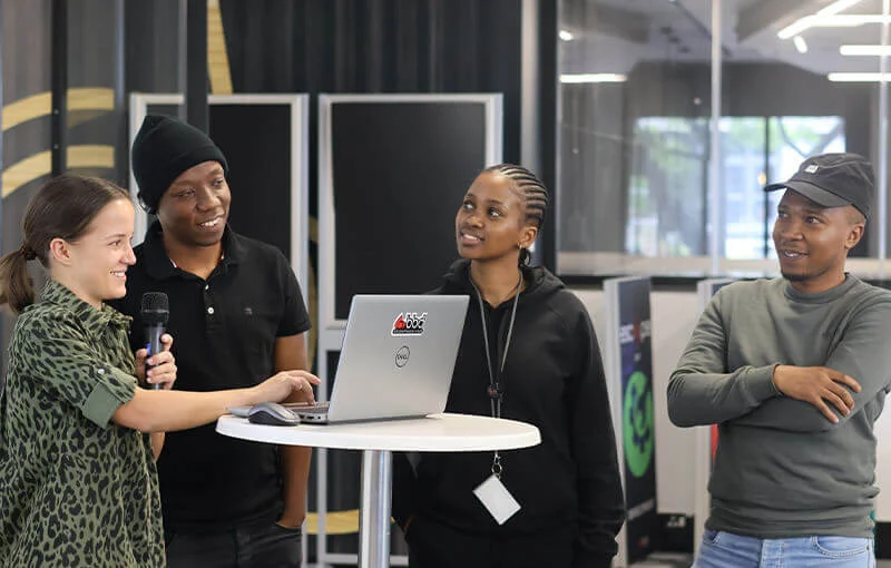 A group of four individuals, smiling and standing around a laptop on a round table. One person speaks into a microphone, creating a collaborative and positive atmosphere.